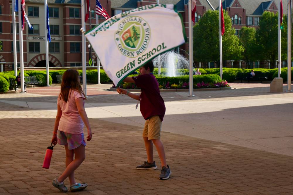 Student waves "Official Michigan Green School" flag in front of a plaza with a fountain